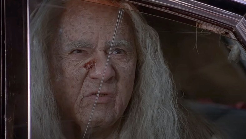 A close up an aging white haired carney with a open sore on his nose, played by Michael Constantine. looks out from a cracked car window. From the film adaptation of Stephen King's Thinner.