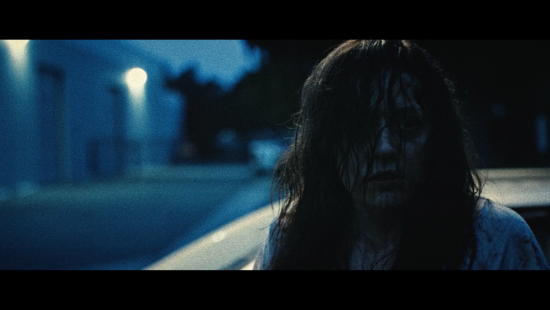 A woman with a blood-spattered face looks dazed as she stands beside a parked car