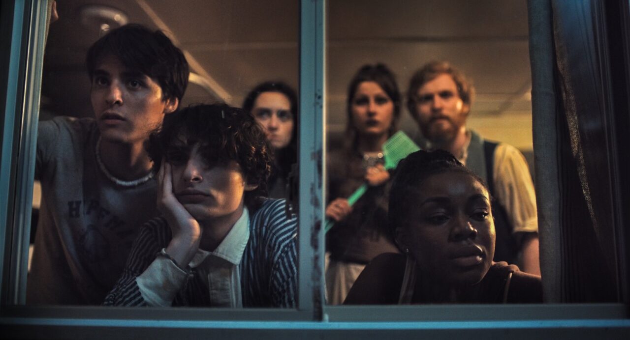 A group of six young people stand in front of a window looking out from the inside of a cabin