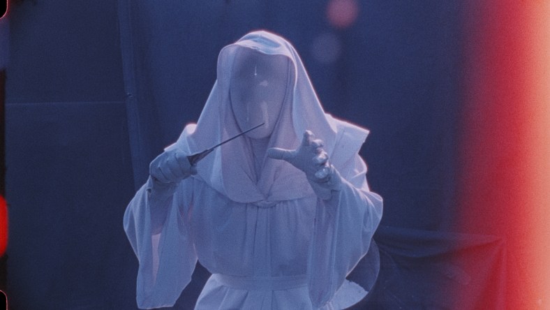 A figure in a white robe, white hood, and white mask that completely obscures their face holds a knife menacingly in one hand and reaches forward with the other.