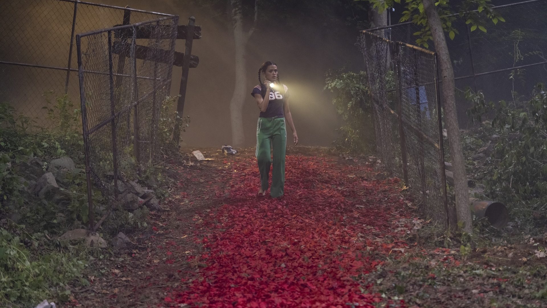 A teenage girl with pigtails holds a flashlight as she walks down a path in a wooded area. The path is lined with red rose petals. 