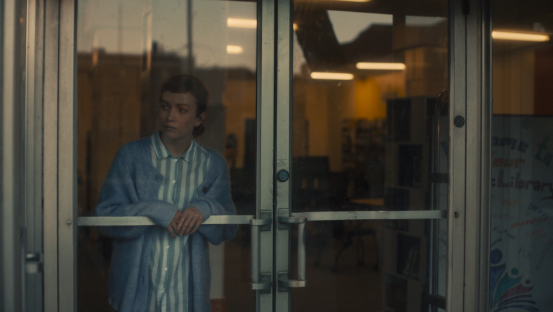 Woman stands on the inside of a glass door of a library in a still from the film Darkest Miriam