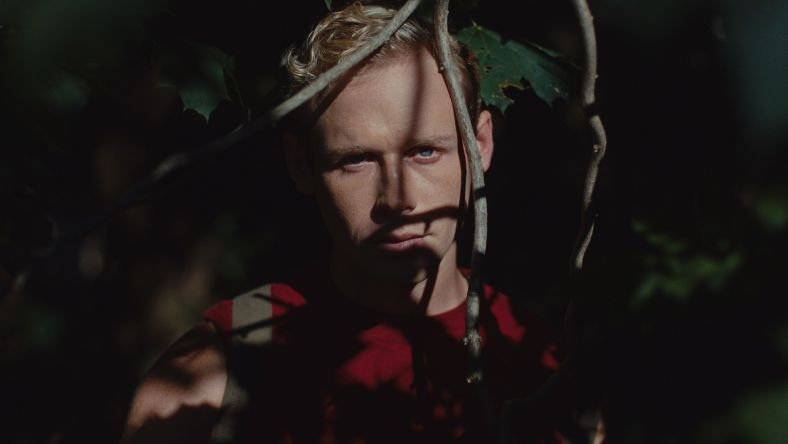 Blonde man stands in a forested area with hanging branches in front of his face in a still from the film Haze